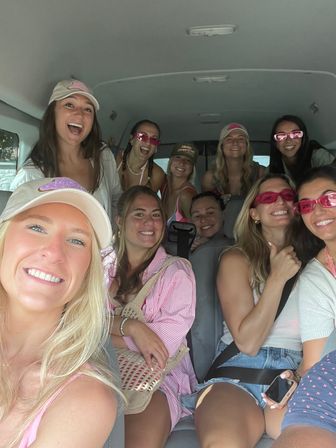 Group selfie of smiling young women in a passenger van wearing summer outfits, baseball caps and pink sunglasses, packed in for a fun road-trip or beach day.