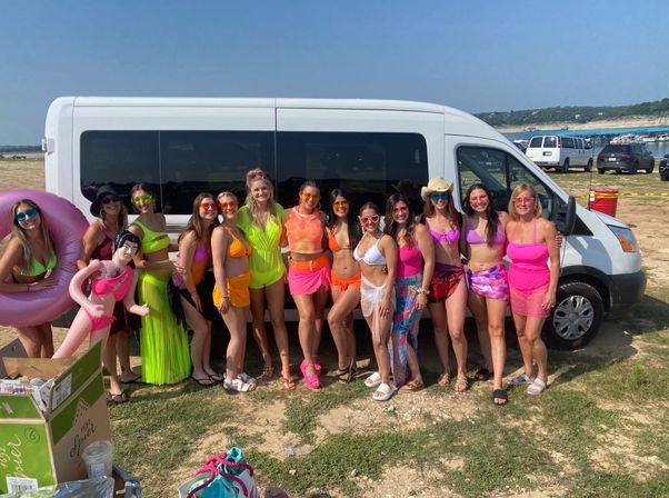 Cheerful group of friends in colorful neon swimsuits posing in front of a white passenger van at a sunny lakeside beach with boats and sandy shoreline in the background.