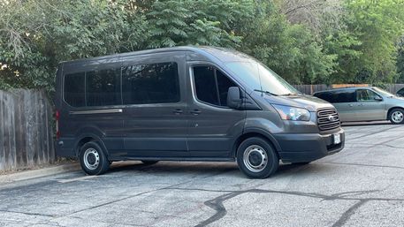 Side view of a large gray passenger van parked on asphalt in a suburban lot beside a wooden fence and leafy trees, with another minivan in the background.