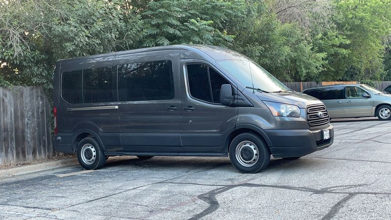 Side view of a large gray passenger van parked on asphalt in a suburban lot beside a wooden fence and leafy trees, with another minivan in the background.