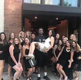 Bachelorette crew — woman in a white dress lifted by a friend outside a downtown bar entrance, surrounded by smiling women in black dresses on the city sidewalk.