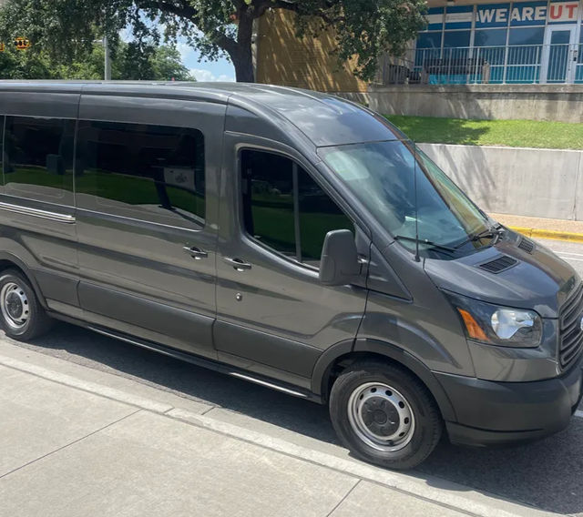 Sleek gray passenger van parked on a sunny urban curb beside a sidewalk and grassy embankment with trees in the background