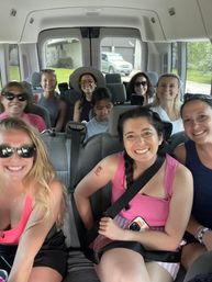 Cheerful group of women riding in a passenger van, smiling in summer clothes with sunglasses and hats, seatbelts fastened, parked in a suburban driveway for a road-trip vibe.