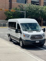 White Ford passenger van parked curbside on a sunny downtown street with a brick office building, traffic lights, sidewalk and shady trees.