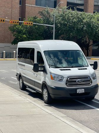 White Ford passenger van parked curbside on a sunny downtown street with a brick office building, traffic lights, sidewalk and shady trees.