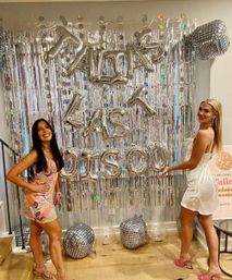 Two smiling women posing at a bachelorette party in Austin, Texas, in front of a silver fringe backdrop with metallic letter balloons and disco-ball balloons on the floor.