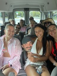 Smiling group of friends in a passenger van on a summer road trip, women in swimwear and sun hats posing together, one holding a novelty face cutout.