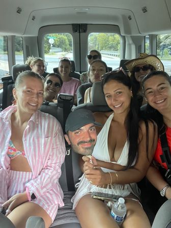 Smiling group of friends in a passenger van on a summer road trip, women in swimwear and sun hats posing together, one holding a novelty face cutout.