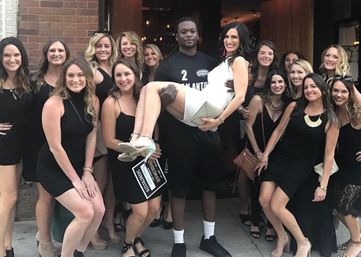 Bachelorette party: woman in white lifted and smiling while friends in black dresses pose outside a downtown bar entrance — lively city nightlife group photo
