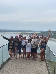 Group of friends in summer outfits and matching pink visors posing on a glass‑railed balcony overlooking a large lake and rolling hills — cheerful lakeside getaway scene.