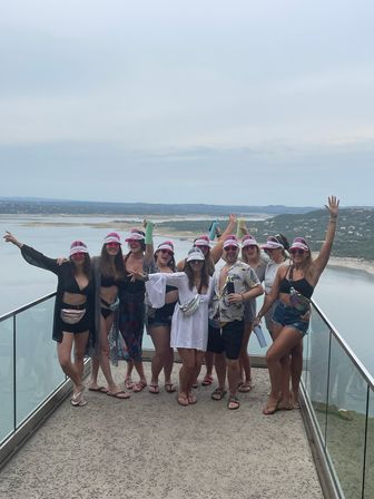 Group of friends in summer outfits and matching pink visors posing on a glass‑railed balcony overlooking a large lake and rolling hills — cheerful lakeside getaway scene.