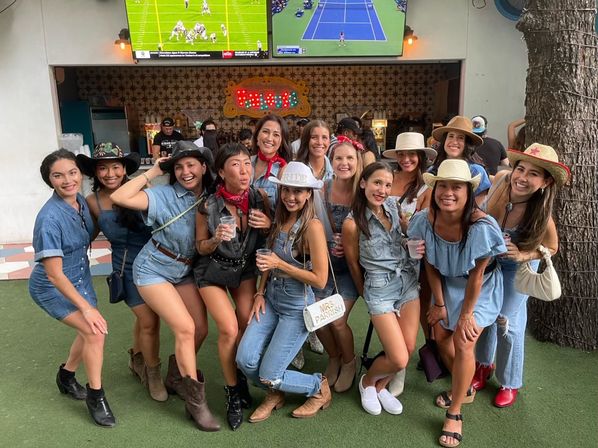 Smiling group of women in denim and cowgirl outfits posing and laughing at an outdoor bar patio, many wearing cowboy hats and boots and holding drinks with large TVs showing sports overhead.