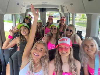 Smiling group of women on a bachelorette party van wearing pink tops, neon 'PARTY' caps and sunglasses, cheerfully waving through suburban streets.