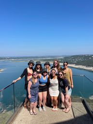 Group of nine adults posing on a glass-railed hilltop overlook under a clear blue sky, smiling in summer outfits and sun hats with a panoramic lake/reservoir and rolling green hills below.