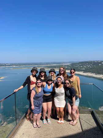 Group of nine adults posing on a glass-railed hilltop overlook under a clear blue sky, smiling in summer outfits and sun hats with a panoramic lake/reservoir and rolling green hills below.