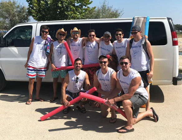 Ten friends in matching white tank tops and swimwear posing with colorful pool noodles in front of a white van on a sunny beach trip.