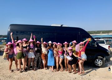 Group of women in pink cowboy hats and summer outfits posing in front of a black party van on a sunny sandy beach with blue sky and coastal water