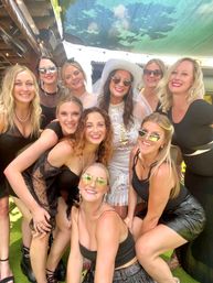 Smiling group of women at a sunny outdoor patio bachelorette party, bride in a white hat and beaded dress surrounded by friends in black outfits and sunglasses, playful celebratory vibe