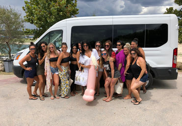 Group of women in swimwear posing in front of a white shuttle van in a coastal parking lot, holding beach bags and a large pink novelty inflatable float under a cloudy sky — fun bachelorette/beach getaway scene.