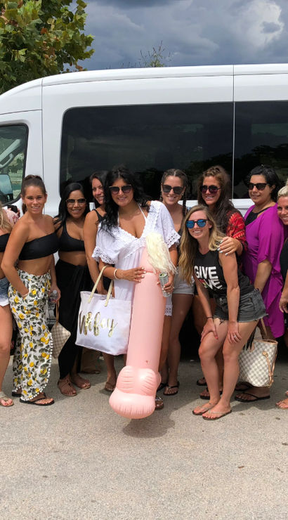 Group of women in swimwear posing in front of a white shuttle van in a coastal parking lot, holding beach bags and a large pink novelty inflatable float under a cloudy sky — fun bachelorette/beach getaway scene.