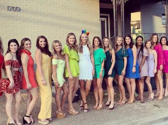 Bachelorette party group photo of women in colorful cocktail dresses lined up outside a light-brick building on a city sidewalk, bride-to-be in white with rainbow headpiece.