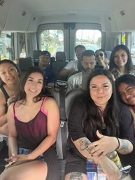 Smiling group selfie inside a passenger van on a sunny suburban road trip — friends in casual summer clothes, visible tattoos, drinks and laughter filling the seats.
