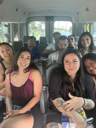 Smiling group selfie inside a passenger van on a sunny suburban road trip — friends in casual summer clothes, visible tattoos, drinks and laughter filling the seats.