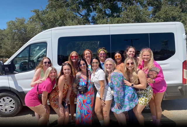 Group of women friends in colorful retro outfits smiling and posing in front of a white passenger van on a sunny outdoor parking lot