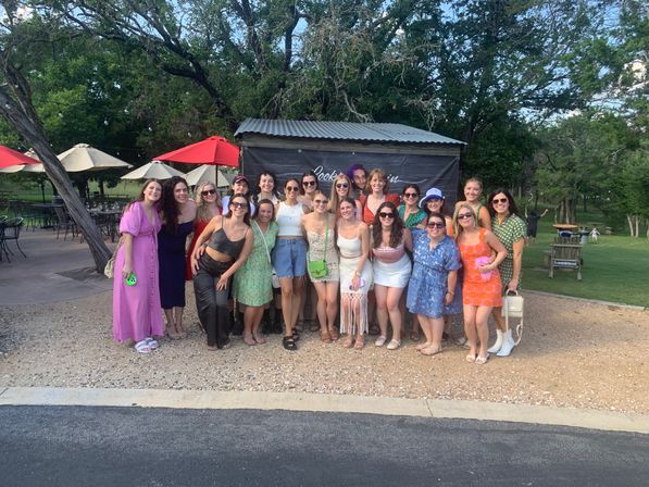 Smiling group of about 20 women in colorful summer outfits posing outdoors at a sunny park-like venue with red patio umbrellas, tables, and trees