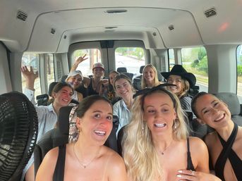 Smiling group of friends inside a passenger van on a daytime city road trip, wearing casual summer outfits and hats with a fun, candid party-shuttle vibe.