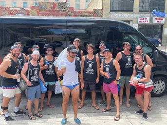 Happy bachelor-party group in matching black tank tops posing with drinks by a black party van on a sunny downtown street with colorful brick storefronts.