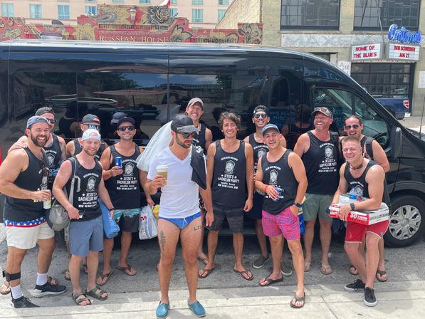 Happy bachelor-party group in matching black tank tops posing with drinks by a black party van on a sunny downtown street with colorful brick storefronts.
