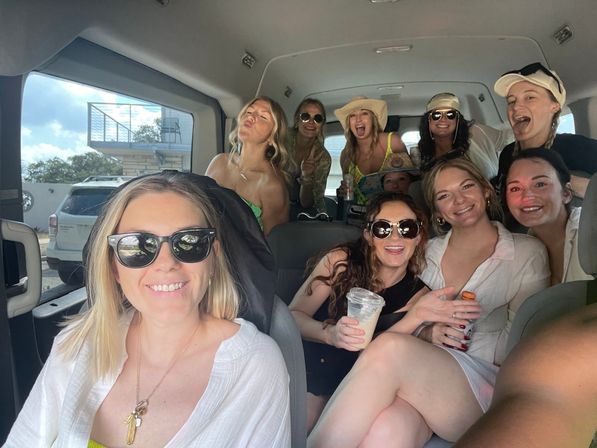 Smiling group of friends packed into a passenger van on a sunny coastal getaway, wearing sunglasses, hats and summer outfits, holding drinks and posing for a cheerful selfie.