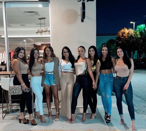 Eight women in stylish evening outfits smiling and posing on a sidewalk outside a modern restaurant at night with palm trees in the background