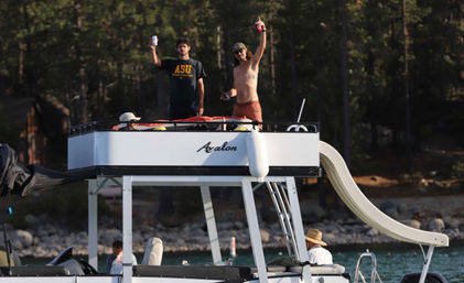 Two men on the upper deck of a pontoon boat raising canned drinks, one shirtless wearing a hat, with a waterslide and ladder, anchored near a pine‑lined lakeshore on a sunny summer day.