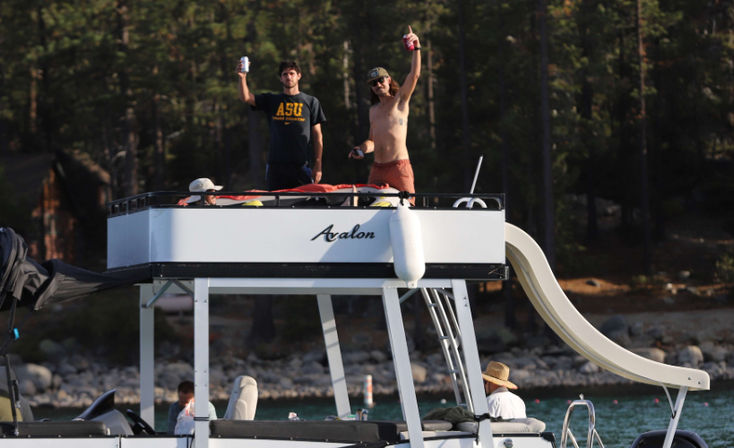 Two men on the upper deck of a pontoon boat raising canned drinks, one shirtless wearing a hat, with a waterslide and ladder, anchored near a pine‑lined lakeshore on a sunny summer day.