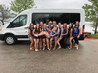 Group of a dozen women in matching red-white-blue tank tops posing and smiling in front of a white passenger van by a lake, one woman held horizontally, trees and cloudy sky in the background