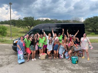 Large group of women in summer clothes posing excitedly by a black passenger van in a roadside parking lot near grassy trees under stormy clouds, holding beach bags and drinks — girls' weekend road trip.