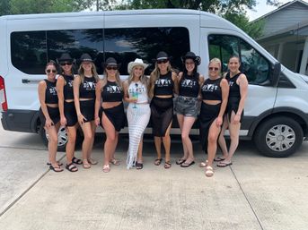 Cowgirl-themed bachelorette party group of ten women in matching black “Howdy” crop tops and cowboy hats, bride in white holding a cocktail, posing in front of a white passenger van on a sunny suburban driveway.
