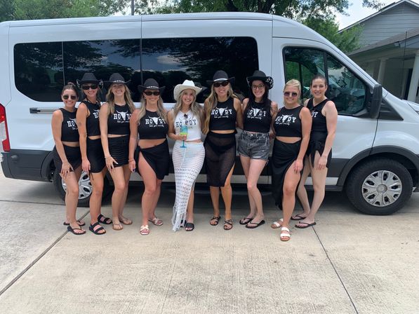 Cowgirl-themed bachelorette party group of ten women in matching black “Howdy” crop tops and cowboy hats, bride in white holding a cocktail, posing in front of a white passenger van on a sunny suburban driveway.