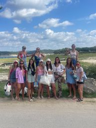 Group of friends at a lakeside overlook under a bright blue sky with fluffy clouds, wearing summer outfits and sunglasses and holding a 'BRIDE' tote and party supplies — fun bachelorette weekend by the water.