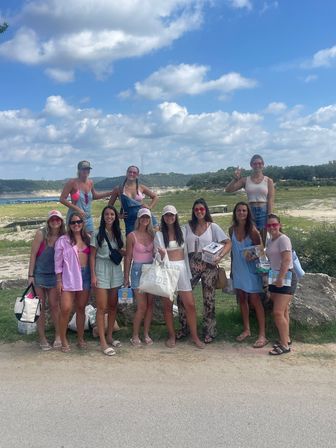 Group of friends at a lakeside overlook under a bright blue sky with fluffy clouds, wearing summer outfits and sunglasses and holding a 'BRIDE' tote and party supplies — fun bachelorette weekend by the water.