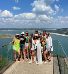 Group of women in summer outfits posing on a glass‑railed cliffside overlook above a blue lake and rolling hills under a bright sky with puffy clouds
