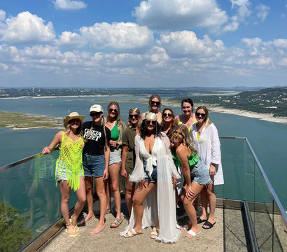 Group of women in summer outfits posing on a glass‑railed cliffside overlook above a blue lake and rolling hills under a bright sky with puffy clouds