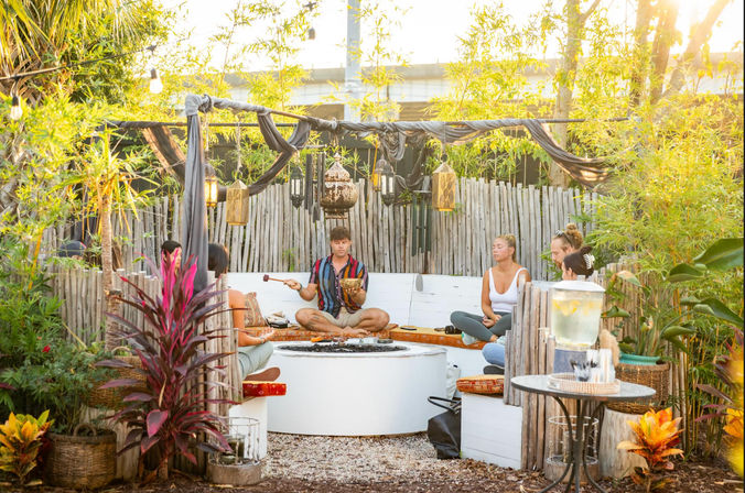 Boho outdoor meditation circle in a tropical garden at sunset — people seated around a white firepit while a leader plays a singing bowl, bamboo fence and hanging lanterns.