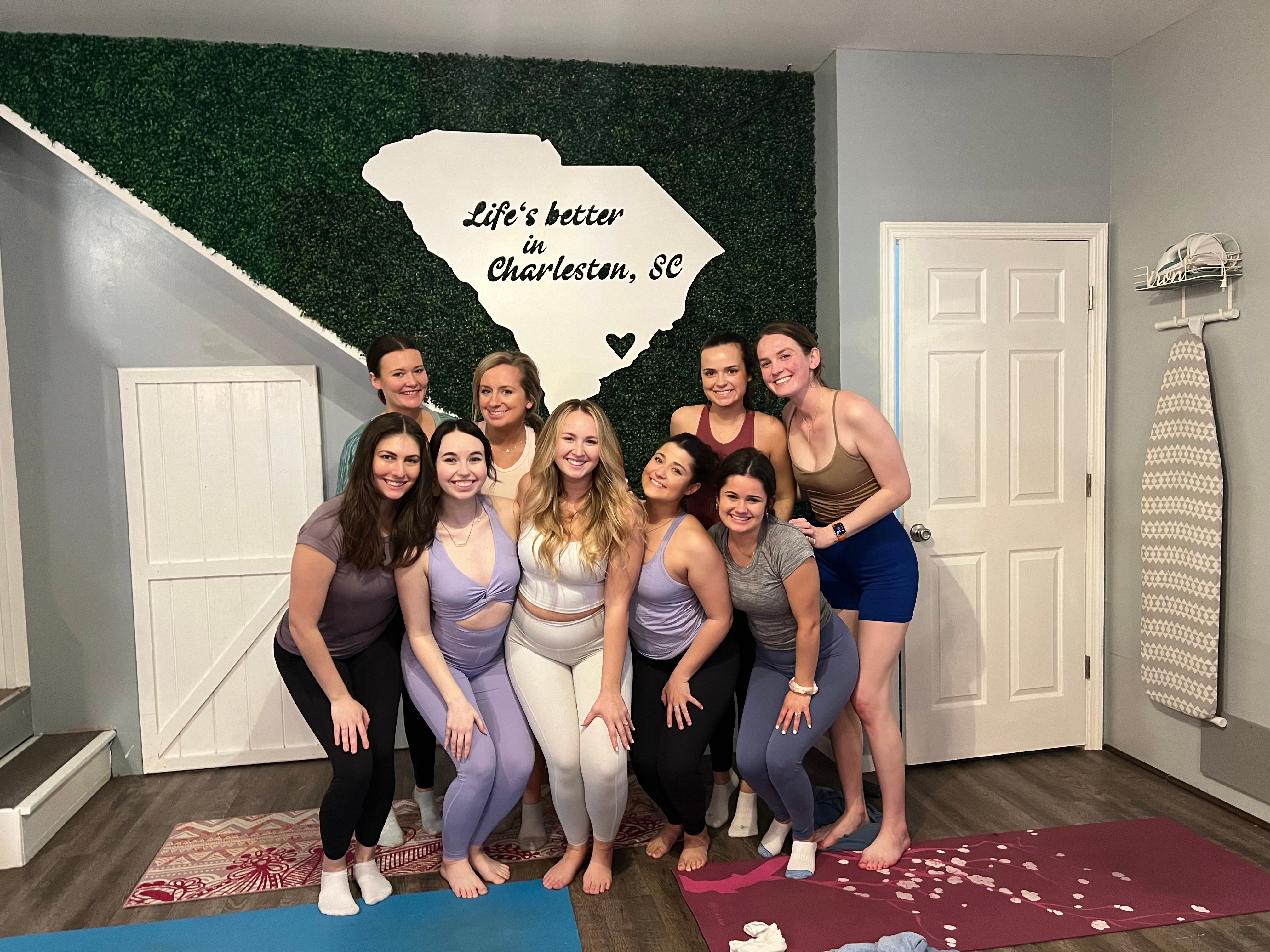 Nine women in colorful yoga attire smiling and posing on mats inside a studio in front of a greenery wall with a white South Carolina silhouette that reads Life's better in Charleston, SC