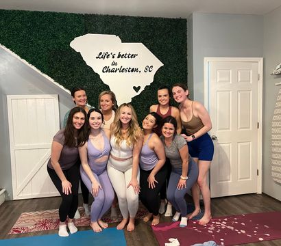 Nine women in colorful yoga attire smiling and posing on mats inside a studio in front of a greenery wall with a white South Carolina silhouette that reads Life's better in Charleston, SC