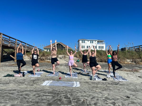 Group of nine people practicing tree pose during a beach yoga session on a sunny sandy shore with coastal houses and a wooden boardwalk under a clear blue sky.