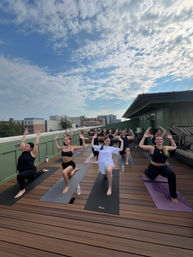 Rooftop yoga class on a wooden deck with a group in low-lunge pose, arms raised, water bottles and city skyline under a partly-cloudy sky.