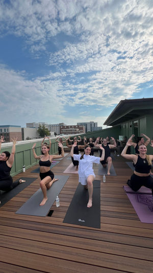Rooftop yoga class on a wooden deck with a group in low-lunge pose, arms raised, water bottles and city skyline under a partly-cloudy sky.
